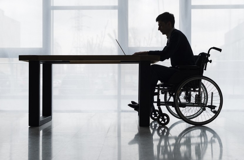 Foto de um homem cadeirante usando um notebook em cima de uma mesa. Ele está sozinho.