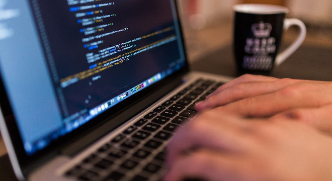 Foto em close de duas mãos digitando no teclado de um notebook. Na tela do equipamento, há uma tela preta com linhas de códigos.